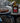 A bottle of pure alaska birch syrup stands on a rustic wood slice next to a plate of grilled salmon with lemon slices, set outdoors on stacked logs with a blue-and-white checkered cloth.