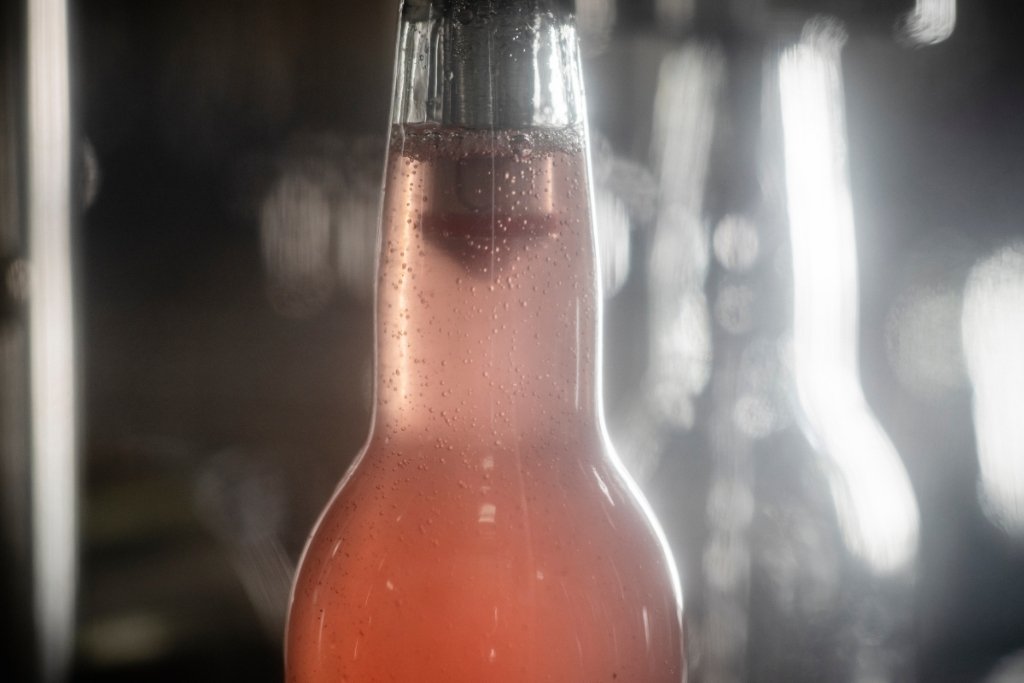 A close-up of a clear glass birch brew fermented birch soda bottle filled with a pinkish-orange carbonated beverage, covered in condensation droplets, set against a dark blurred background with bright light flares.