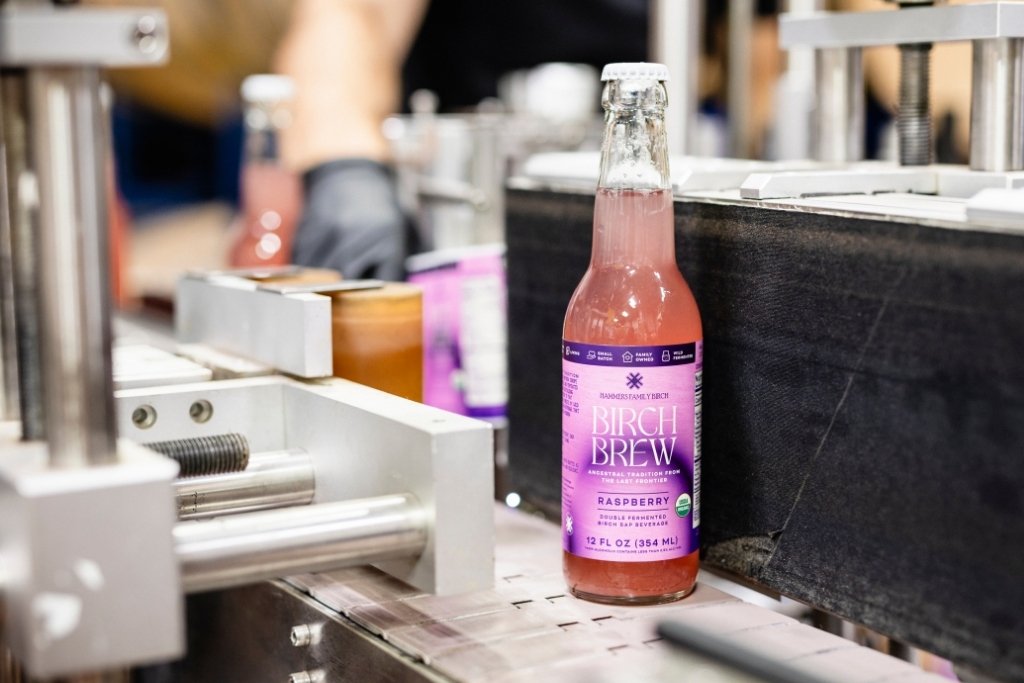 A bottle of Birch Brew Raspberry sparkling fermented birch sap drink sits on a conveyor belt in a production setting, with machinery and another bottle blurred in the background.