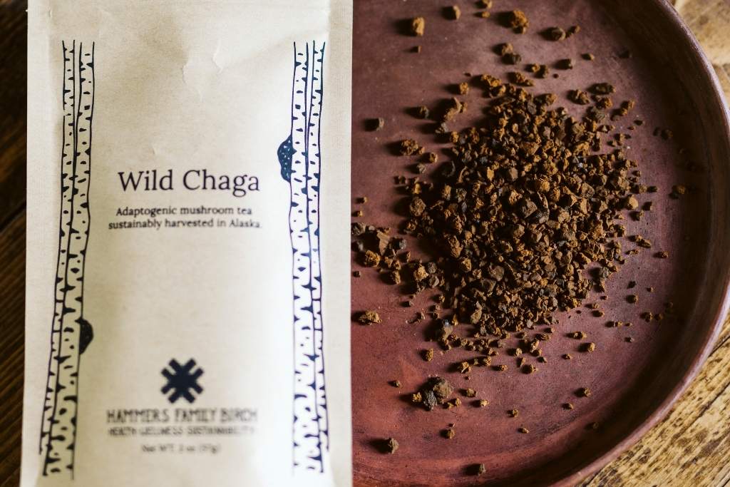 A package of Hammer's Family Birch Wild Chaga mushroom tea stands next to a dark brown plate filled with loose dark brown chaga pieces on a wooden surface.